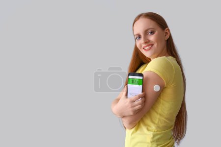 Woman with glucose sensor using mobile phone for measuring of blood sugar level on grey background