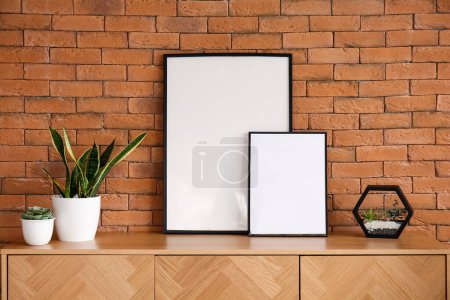 Téléchargez les photos : Élégante armoire en bois avec cadres vierges et plantes d'intérieur près du mur de briques brunes - en image libre de droit
