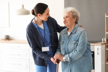 Young caregiver helping senior woman with walking stick in kitchen