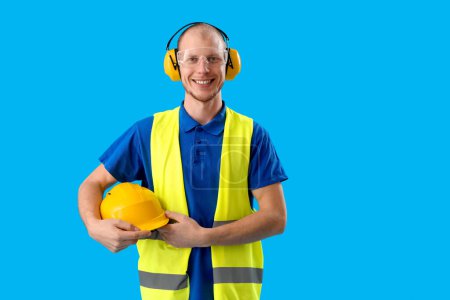 Foto de Hombre trabajador en chaleco, con casco y auriculares protectores sobre fondo azul - Imagen libre de derechos