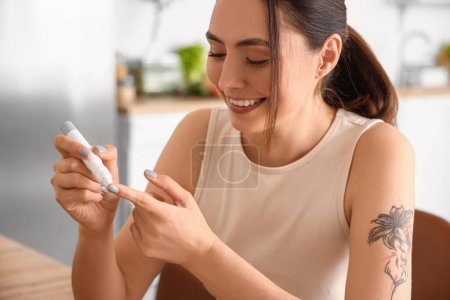 Photo for Young diabetic woman measuring blood sugar level with lancet pen in kitchen, closeup - Royalty Free Image
