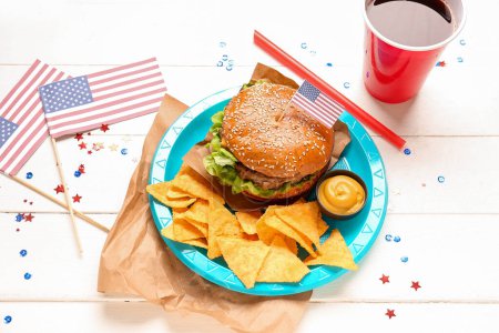 Composition with tasty burger, nachos, sauce, cola and paper American flags on light wooden background. Memorial Day celebration
