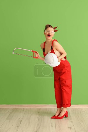 Téléchargez les photos : Jeune femme pin-up en uniforme rouge avec scie à métaux et casque près du mur vert - en image libre de droit