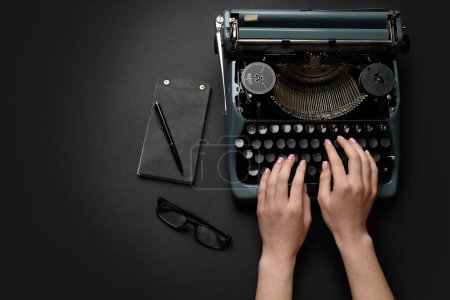 Foto de Mujer escribiendo en máquina de escribir con anteojos, cuaderno y pluma sobre fondo negro - Imagen libre de derechos