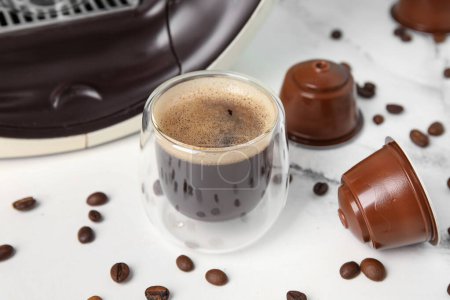 Glass of delicious espresso, capsule coffee maker, pods and beans on white table