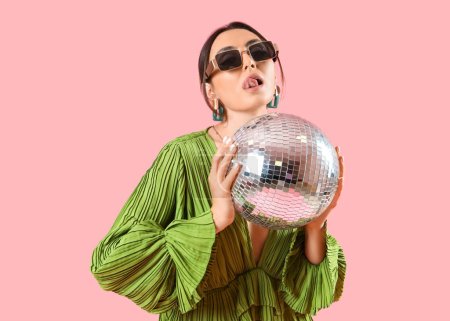 Téléchargez les photos : Belle femme à la mode en robe verte et lunettes de soleil avec boule disco posant sur fond rose - en image libre de droit