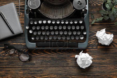 Vintage typewriter with notebook, eyeglasses and houseplant on brown wooden background