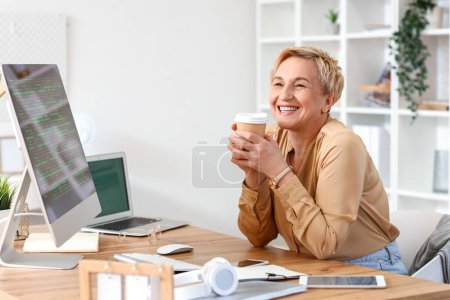 Téléchargez les photos : Femme mature programmeuse avec tasse de café travaillant à la table dans le bureau - en image libre de droit