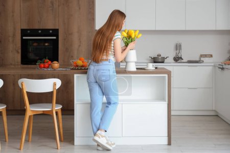 Téléchargez les photos : Belle jeune femme sentant fleurs de tulipes en fleurs dans la cuisine moderne - en image libre de droit