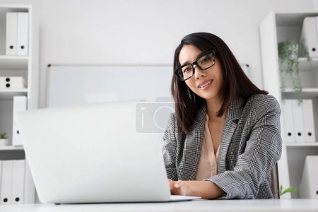 Téléchargez les photos : Belle femme d'affaires asiatique dans des lunettes élégantes de travail avec ordinateur portable au bureau - en image libre de droit