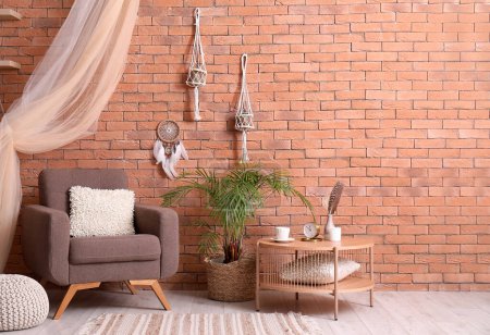 Interior of modern living room with armchair, table, plant and dream catcher hanging on brick wall-stock-photo