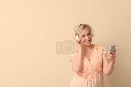 Mature woman in headphones with mobile phone on beige background