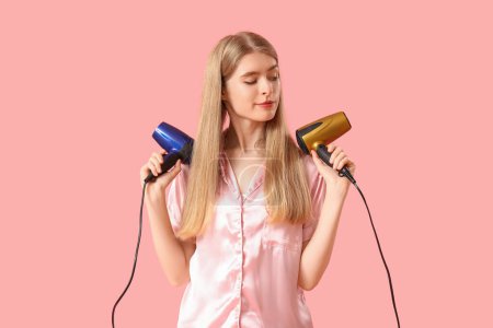 Photo for Thoughtful young blonde woman in pajamas with hair dryers on pink background - Royalty Free Image
