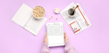 Foto de Manos femeninas sosteniendo la tableta de ordenador, calendario, periódico y desayuno sobre fondo lila - Imagen libre de derechos