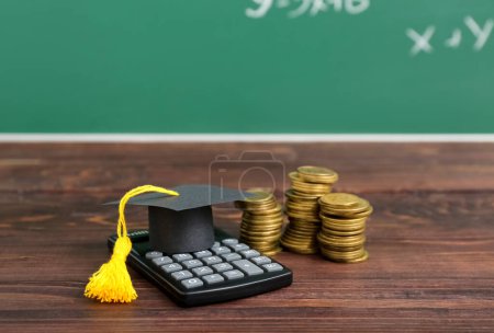 Foto de Tapa de graduación con calculadora y pilas de monedas en mesa de madera marrón contra pizarra. Concepto de préstamo estudiantil - Imagen libre de derechos
