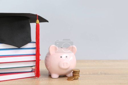 Piggy bank, stack of coins, books and graduation hat on wooden table against grey background. Student loan concept