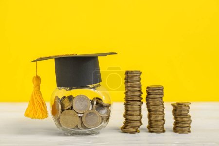 Foto de Tarro con dinero, gorra de graduación y pilas de monedas sobre mesa de madera sobre fondo amarillo. Concepto de préstamo estudiantil - Imagen libre de derechos