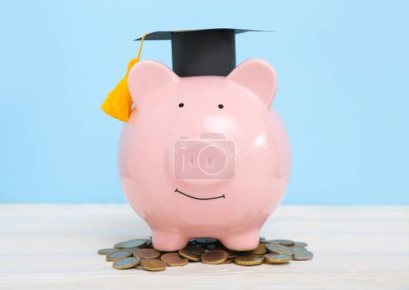 Foto de Hucha de cerdito en gorra de graduación y monedas en mesa de madera blanca sobre fondo azul. Concepto de préstamo estudiantil - Imagen libre de derechos