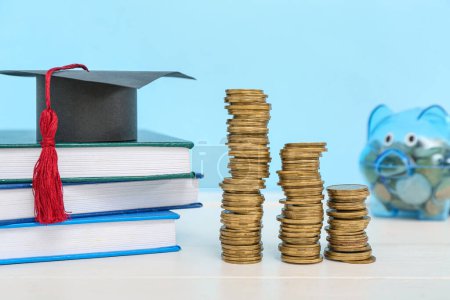 Foto de Montones de monedas, libros, gorra de graduación y alcancía sobre una mesa blanca sobre fondo azul. Concepto de préstamo estudiantil - Imagen libre de derechos
