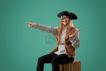 Foto de Hermosa pirata femenina con spyglass sentado en la caja de madera y apuntando a algo contra el fondo verde - Imagen libre de derechos