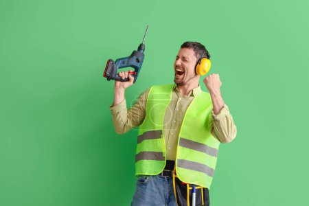 Foto de Constructor masculino en protectores auditivos con taladro sobre fondo verde - Imagen libre de derechos