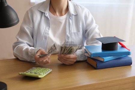 Foto de Estudiante sentada en la mesa con libros y calculadora y contando dinero. Concepto de préstamo estudiantil - Imagen libre de derechos