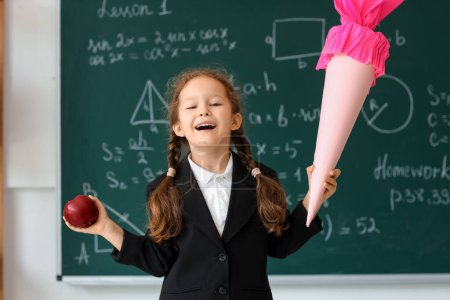 Happy little girl with pink school cone and fresh apple in classroom near blackboard