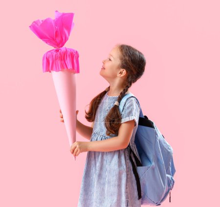 Glückliches kleines Mädchen mit Rucksack und Schultüte auf rosa Hintergrund