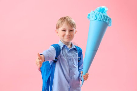Glücklicher kleiner Junge mit blauer Schultüte mit Daumen-hoch-Geste auf rosa Hintergrund