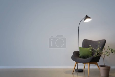 Glowing lamp, grey armchair with cushion and houseplant near grey wall