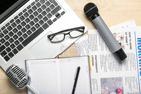 Laptop with eyeglasses, microphones, notebook and newspapers on light wooden background