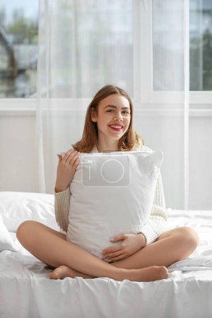 Foto de Hermosa joven en medias beige y con almohada sentada en la cama - Imagen libre de derechos