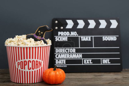 Bucket with tasty popcorn, cookie, pumpkin and clapperboard on wooden table. Halloween celebration
