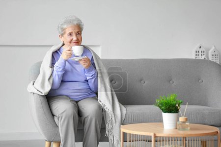 Starsza kobieta z kratą pije kawę na kanapie w domu