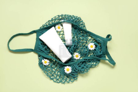 Téléchargez les photos : Sac en maille avec de belles fleurs de camomille et des tubes de produits cosmétiques sur fond vert - en image libre de droit