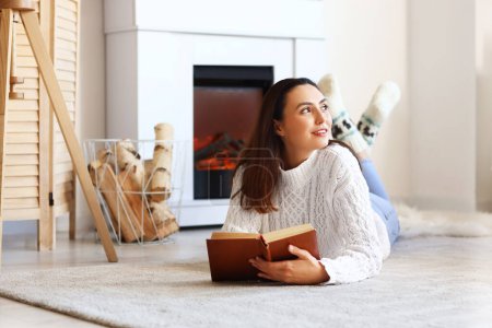 Mujer joven leyendo libro cerca de la chimenea en casa