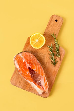 Téléchargez les photos : Panneau en bois de steak de saumon frais dans une pellicule alimentaire en plastique, citron et romarin sur fond jaune - en image libre de droit