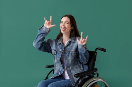 Téléchargez les photos : Jeune femme en fauteuil roulant montrant "cornes du diable" sur fond vert - en image libre de droit