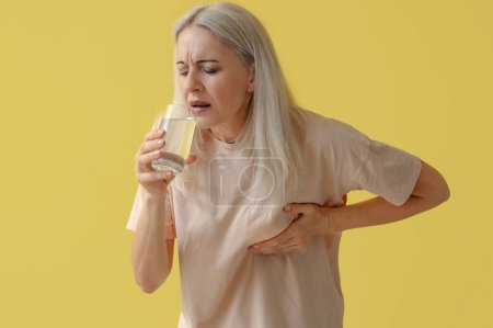 Téléchargez les photos : Femme mûre avec verre d'eau ayant une crise cardiaque sur fond jaune - en image libre de droit