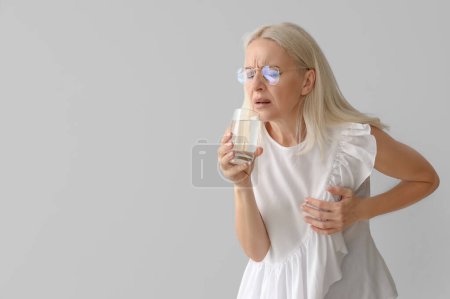 Téléchargez les photos : Femme mûre avec verre d'eau ayant une crise cardiaque sur fond gris - en image libre de droit