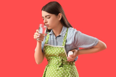 Téléchargez les photos : Jeune femme au foyer avec verre d'eau et bouteille de pilule ayant une crise cardiaque sur fond rouge - en image libre de droit