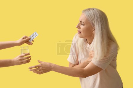 Téléchargez les photos : Femme mature avec crise cardiaque recevant des pilules et un verre d'eau sur fond jaune - en image libre de droit