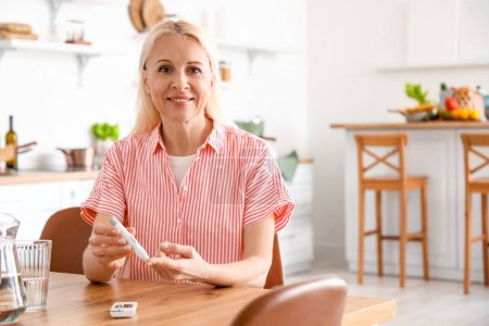 Photo for Mature diabetic woman measuring blood sugar level with lancet pen in kitchen - Royalty Free Image