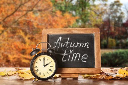 Photo for Alarm clock, chalkboard with text AUTUMN TIME and leaves on table outdoors - Royalty Free Image