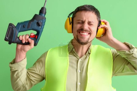 Foto de Constructor masculino en protectores auditivos con taladro sobre fondo verde - Imagen libre de derechos