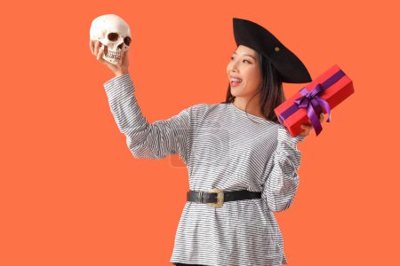 Junge Asiatin als Piratin verkleidet mit Halloween-Geschenk und Totenkopf auf orangefarbenem Hintergrund