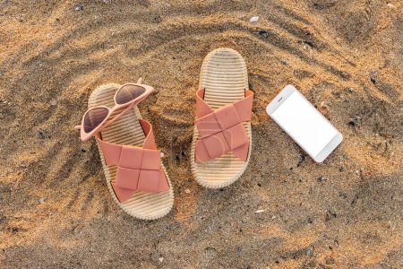 Foto de Chanclas con estilo con gafas de sol y teléfono móvil en la arena en el resort - Imagen libre de derechos