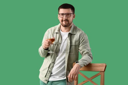 Photo for Young sommelier with glass of wine leaning on wooden chair against green background - Royalty Free Image