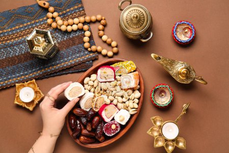 Photo for Female hand with diya lamps, candles, beads and plate of different treats on brown background. Divaly celebration - Royalty Free Image