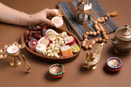 Photo for Female hand with diya lamps, candles, beads and plate of different treats on brown background. Divaly celebration - Royalty Free Image